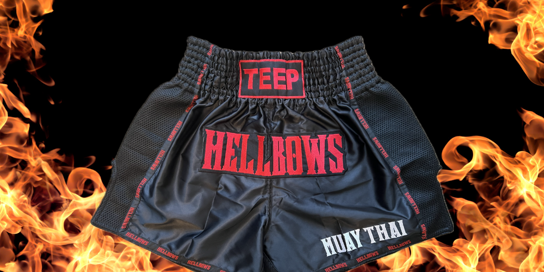 Picture of Black Muay Thai shorts with the word HELLBOWS in red stitched on the front. The word TEEP is stitched in red on the waistband and the words MUAY THAI is stitched in white on the bottom left corner of the shorts. The background to this picture is yellow flames