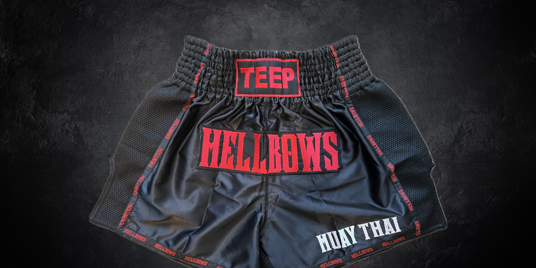 Picture of Black Muay Thai shorts with the word HELLBOWS in red stitched on the front. The word TEEP is stitched in red on the waistband and the words MUAY THAI is stitched in white on the bottom left corner of the shorts. The shorts sit on a black background